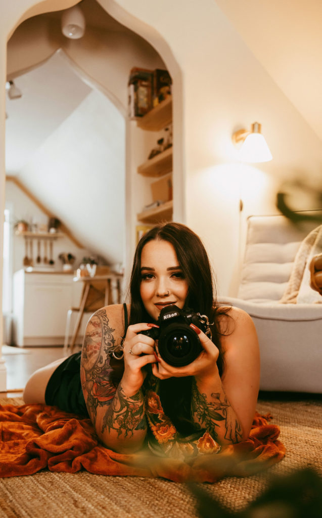 Sharlie Faye lays on the floor of a boho apartment holding her camera, smiling. 